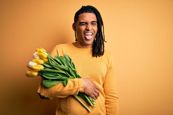 Young african american afro romantic man with dreadlocks holding bouquet of yellow tulips winking looking at the camera with sexy expression, cheerful and happy face. - Фото, изображение
