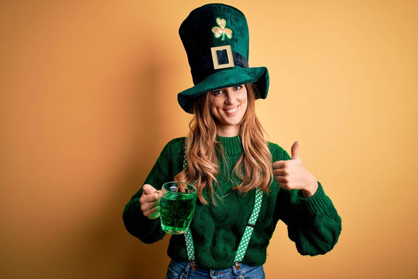 Beautiful woman wearing hat drinking jar of green beverage celebrating saint patricks day happy with big smile doing ok sign, thumb up with fingers, excellent sign - Foto, imagen