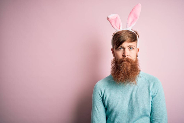 Ирландский хипстер с бородой в ушах пасхального кролика на изолированном розовом фоне с серьезным выражением лица. Простой и естественный взгляд на камеру
. - Фото, изображение