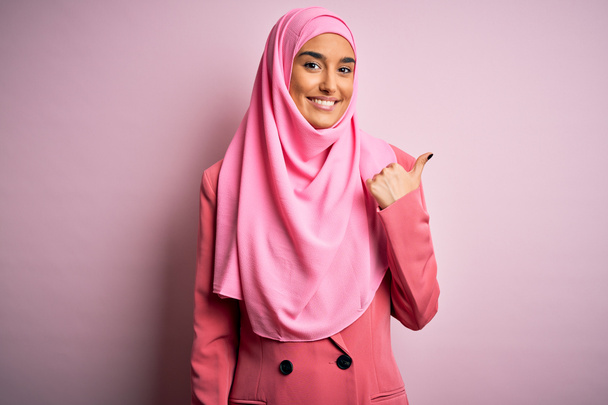 Νεαρή όμορφη μελαχρινή επιχειρηματίας που φοράει ροζ muslim χιτζάμπ και επαγγελματικό μπουφάν χαμογελώντας με χαρούμενο πρόσωπο και δείχνοντας στο πλάι με τον αντίχειρα προς τα πάνω. - Φωτογραφία, εικόνα