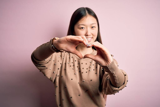 Młoda piękna azjatycka kobieta nosi modę i elegancki sweter na różowym tle solated uśmiechając się w miłości robi kształt symbolu serca z rąk. Romantyczna koncepcja. - Zdjęcie, obraz