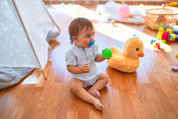 Criança adorável sentado no chão usando chupeta em torno de lotes de brinquedos no jardim de infância
 - Foto, Imagem