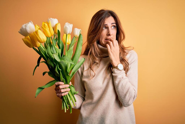 Młoda piękna brunetka trzyma bukiet żółtych tulipanów na odizolowanym tle wyglądając zestresowany i nerwowy z rękami na ustach obgryzając paznokcie. Problem lękowy. - Zdjęcie, obraz