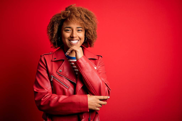 Joven mujer afro afroamericana hermosa con el pelo rizado usando chaqueta roja casual mirando confiado en la cámara sonriendo con los brazos cruzados y la mano levantada en la barbilla. Pensando positivamente
. - Foto, imagen
