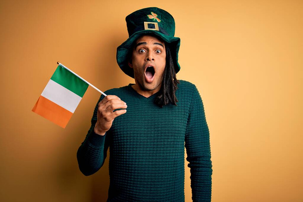 Africký Američan v zeleném klobouku drží irskou irskou irskou vlajku oslavující svaté patricks den vyděšený v šoku s překvapenou tváří, strach a vzrušení výrazem strachu - Fotografie, Obrázek