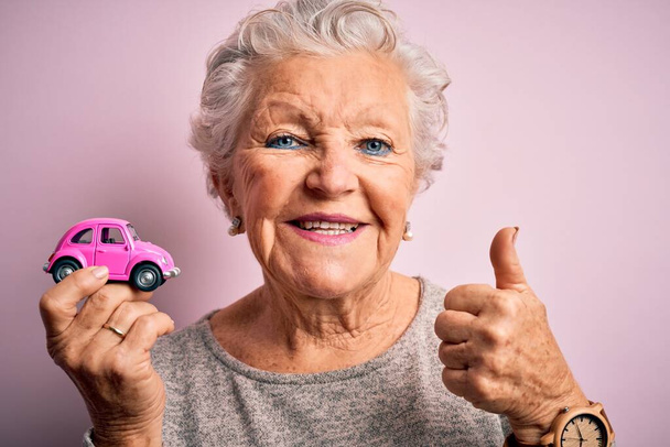 Senior bella donna che tiene piccola auto in piedi sopra isolato sfondo rosa felice con grande sorriso facendo segno ok, pollice in su con le dita, segno eccellente
 - Foto, immagini