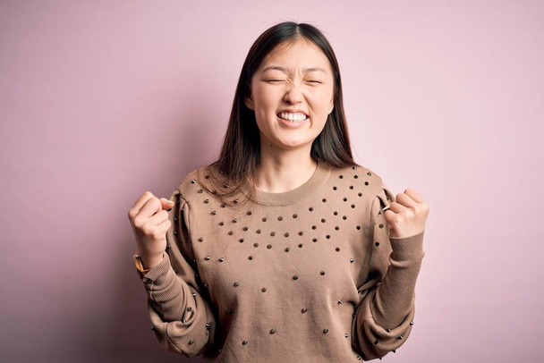 junge schöne asiatische Frau trägt Mode und eleganten Pullover über rosa solated Hintergrund sehr glücklich und aufgeregt dabei Siegergeste mit erhobenen Armen, lächelnd und schreiend nach Erfolg. Festkonzept. - Foto, Bild