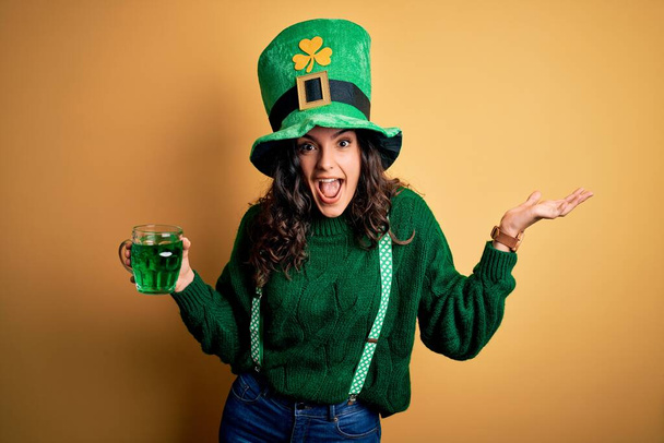 Beautiful curly hair woman wearing hat drinking jar of green beverage on saint patricks day very happy and excited, winner expression celebrating victory screaming with big smile and raised hands - Fotó, kép