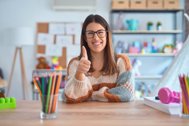 Young beautiful teacher woman wearing sweater and glasses sitting on desk at kindergarten doing happy thumbs up gesture with hand. Approving expression looking at the camera showing success. - Foto, immagini