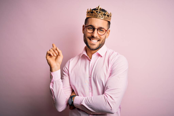 Jonge knappe zakenman draagt gouden kroon als koning over roze achtergrond met een grote glimlach op het gezicht, wijzend met hand en vinger naar de zijkant kijkend naar de camera. - Foto, afbeelding