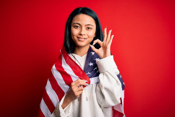 junge schöne Chinesin trägt Vereinigte Staaten Flagge über isoliertem roten Hintergrund tun ok Zeichen mit den Fingern, ausgezeichnetes Symbol - Foto, Bild