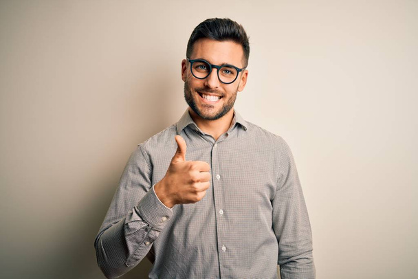 Young handsome man wearing elegant shirt and glasses over isolated white background doing happy thumbs up gesture with hand. Approving expression looking at the camera showing success. - Fotó, kép