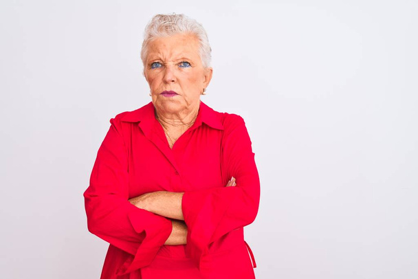 İzole edilmiş beyaz arka plan üzerinde kırmızı bir gömlek giyen yaşlı gri saçlı kadın şüpheci ve gergin, yüzünde çapraz kollu tasvip etmeyen bir ifade var. Negatif kişi. - Fotoğraf, Görsel