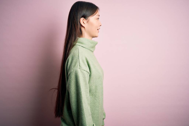 Jovem bela mulher asiática vestindo camisola verde inverno sobre rosa solado fundo olhando para o lado, relaxar perfil posar com rosto natural e sorriso confiante
. - Foto, Imagem