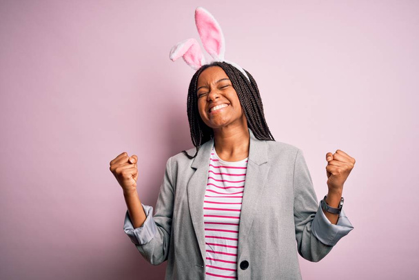 Młoda afrykańska dziewczyna nosi słodkie uszy króliczka wielkanocnego na różowym tle bardzo szczęśliwy i podekscytowany robi gest zwycięzca z podniesionymi ramionami, uśmiech i krzyk o sukces. Koncepcja uroczystości. - Zdjęcie, obraz