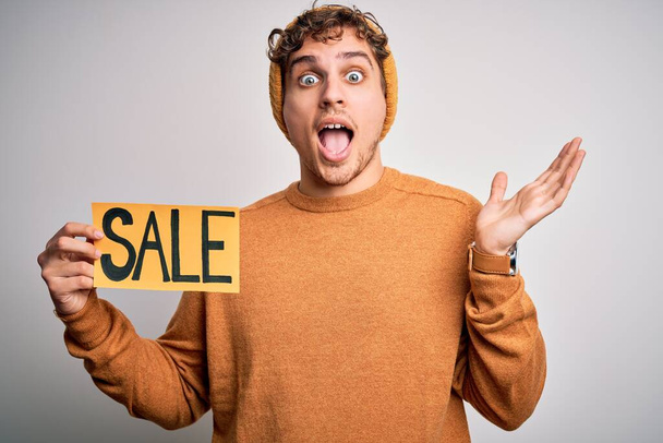 Young blond man with curly hair holding sale poster standing over isolated white background very happy and excited, winner expression celebrating victory screaming with big smile and raised hands - Fotoğraf, Görsel