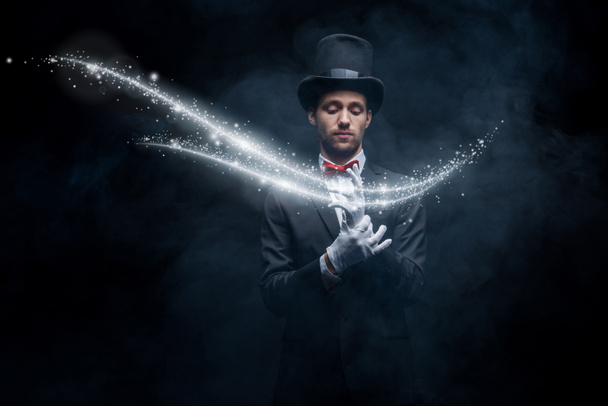 Zauberer in Anzug und Mütze mit Handschuhen in dunklem, verrauchtem Raum mit glühender Illustration - Foto, Bild