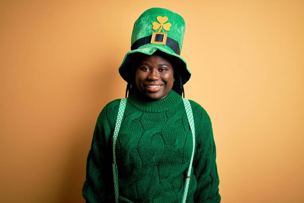 Mujer afroamericana de talla grande con trenzas con sombrero verde y trébol en el día de San Patricio con una sonrisa alegre y fresca en la cara. Persona afortunada
. - Foto, Imagen