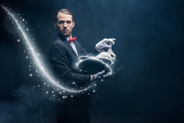 professioneller Zauberer im Anzug zeigt Trick mit weißem Kaninchen im Hut, dunkler Raum mit Rauch und glühender Illustration - Foto, Bild