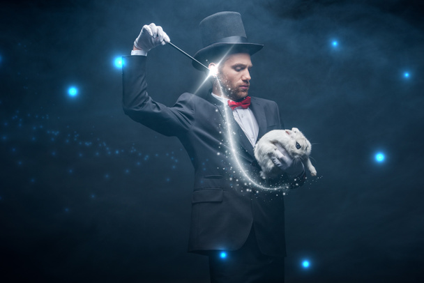 emocionální kouzelník v obleku a klobouku ukazující trik s hůlkou a bílým králíkem, tmavý pokoj s kouřem a zářící ilustrací - Fotografie, Obrázek