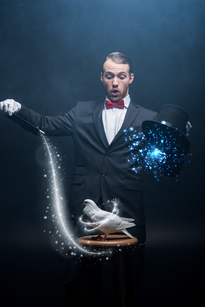 Schockierter Zauberer zeigt Trick mit Taube, Zauberstab und Hut in dunklem Raum mit Rauch und glühender Illustration - Foto, Bild