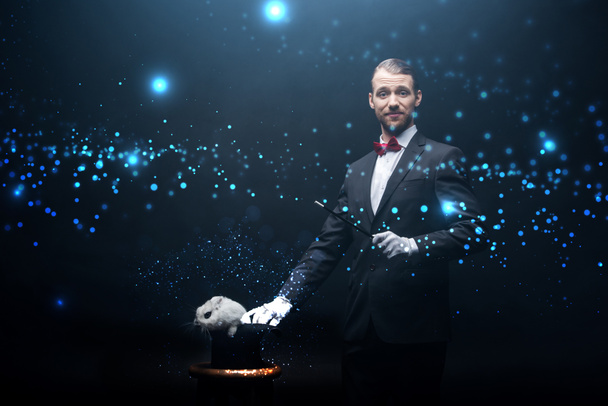 счастливый фокусник в костюме, показывающий фокус с палочкой и белым кроликом в шляпе, темной комнатой с дымом и светящимися иллюстрациями
 - Фото, изображение