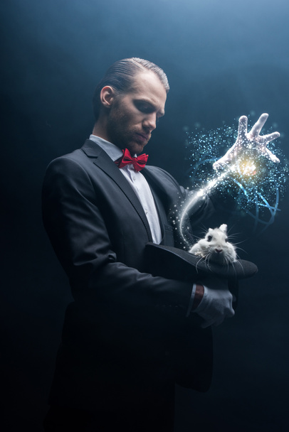 professional magician gesturing over white rabbit in hat, dark room with smoke and glowing illustration - Photo, Image