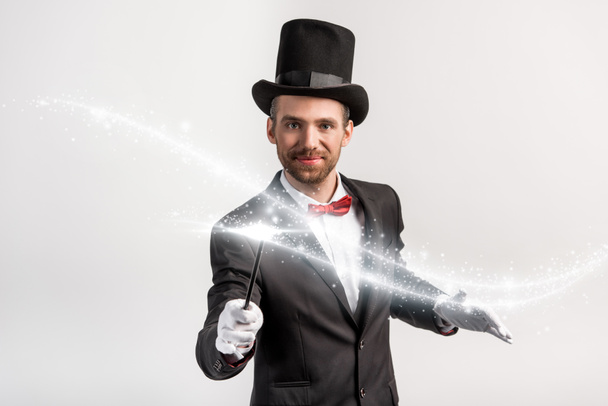 pozitivní kouzelník v obleku a klobouku drží hůlku izolovanou na šedé se zářivou ilustrací - Fotografie, Obrázek