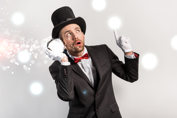 professional magician pointing up and listening to magic ball isolated on grey with glowing illustration - Photo, Image