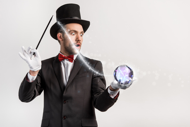 Потрясенный фокусник держит волшебную палочку и волшебный шар, изолированный на сером с яркой иллюстрацией
 - Фото, изображение