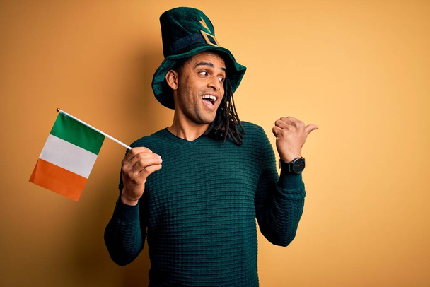 Homme afro-américain portant un chapeau vert tenant le drapeau irlandais célébrant saint patricks jour pointant et montrant avec le pouce sur le côté avec le visage heureux souriant
 - Photo, image
