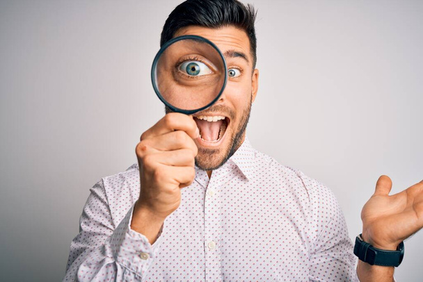 Young detective man looking through magnifying glass over isolated background very happy and excited, winner expression celebrating victory screaming with big smile and raised hands - Foto, Imagen