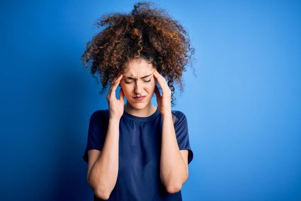 Молодая красивая женщина с вьющимися волосами и пирсинг носить случайные синие футболки с рукой на голове от боли в голове, потому что стресс. Страдания от мигрени
. - Фото, изображение