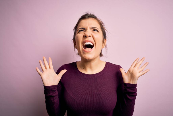 Beautiful young woman wearing casual bun hairstyle over pink isolated background crazy and mad shouting and yelling with aggressive expression and arms raised. Frustration concept. - Photo, Image