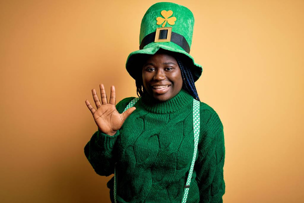 Más tamaño mujer afroamericana con trenzas con sombrero verde con trébol en st patricks día mostrando y apuntando hacia arriba con los dedos número cinco mientras sonríe confiado y feliz
. - Foto, imagen