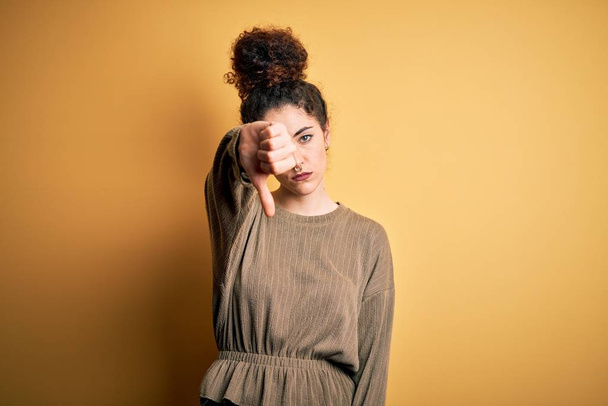Young beautiful brunette woman with curly hair and piercing wearing casual dress looking unhappy and angry showing rejection and negative with thumbs down gesture. Bad expression. - Foto, afbeelding