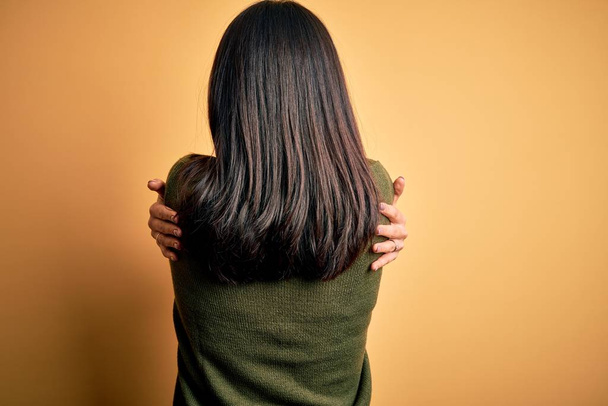 Young brunette woman with blue eyes wearing green casual sweater over yellow background Hugging oneself happy and positive from backwards. Self love and self care - Photo, Image