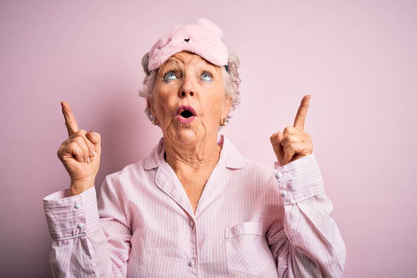 Senior bela mulher vestindo máscara de sono e pijama sobre fundo rosa isolado espantado e surpreso olhando para cima e apontando com os dedos e braços levantados
. - Foto, Imagem