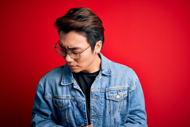 Young handsome chinese man wearing denim jacket and glasses over red background with hand on stomach because indigestion, painful illness feeling unwell. Ache concept. - Φωτογραφία, εικόνα