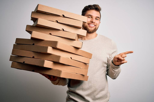 Молодой блондин с бородой и голубыми глазами, держащий картонки пиццы на белом фоне, очень счастлив, указывая рукой и пальцем в сторону.
 - Фото, изображение