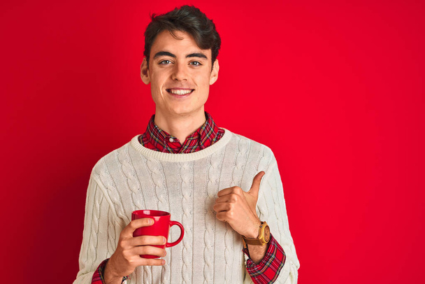 Nastolatek w zimowym swetrze pijący filiżankę kawy na odizolowanym czerwonym tle szczęśliwy z dużym uśmiechem robi znak ok, kciuk do góry palcami, doskonały znak - Zdjęcie, obraz