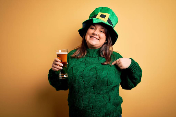 Young beautiful plus size woman wearing green hat celebrating st patricks day drinking beer with surprise face pointing finger to himself - Photo, Image