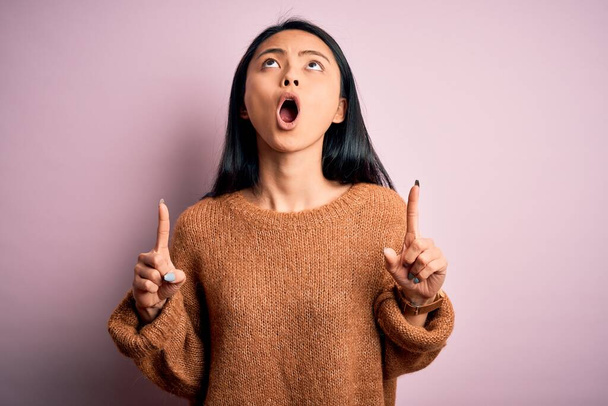 Young beautiful chinese woman wearing casual sweater over isolated pink background amazed and surprised looking up and pointing with fingers and raised arms. - Foto, immagini