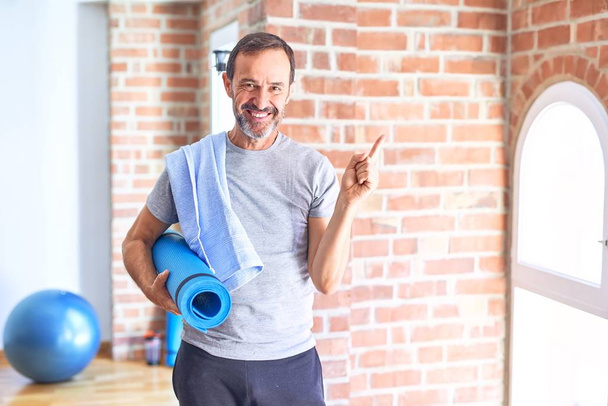 Middle age handsome sportman holding mat and towel standing before do exercise at gym very happy pointing with hand and finger to the side - Photo, Image
