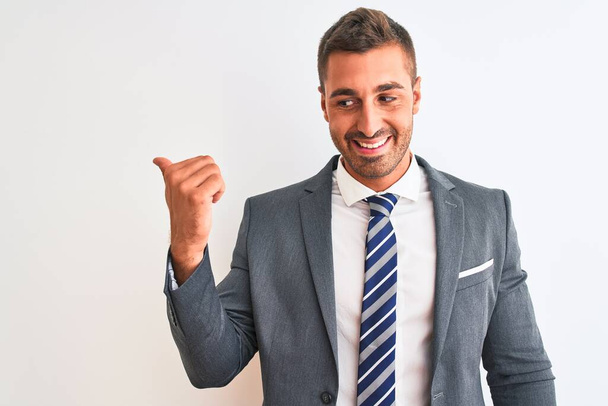 Young handsome business man wearing suit and tie over isolated background smiling with happy face looking and pointing to the side with thumb up. - Valokuva, kuva
