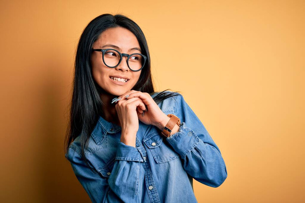 Young beautiful chinese woman wearing casual denim shirt over isolated yellow background laughing nervous and excited with hands on chin looking to the side - 写真・画像