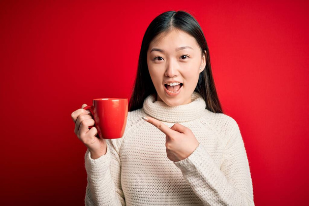 Genç Asyalı kadın izole edilmiş kırmızı arka planda bir fincan sıcak kahve içiyor. El ve parmağıyla işaret ederek çok mutlu. - Fotoğraf, Görsel