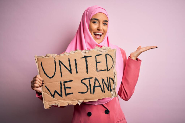 Jonge activistische vrouw draagt roze moslim hijab houden spandoek met verenigde wij staan boodschap zeer gelukkig en opgewonden, winnaar uitdrukking vieren overwinning schreeuwen met grote glimlach en opgeheven handen - Foto, afbeelding
