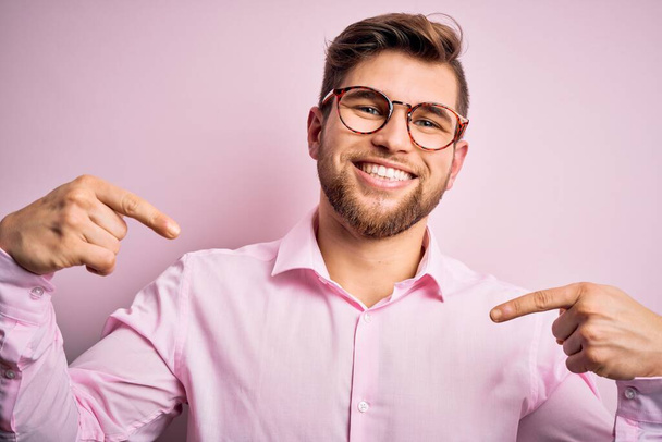 Jonge knappe blonde man met baard en blauwe ogen met roze hemd en bril die er zelfverzekerd uitziet met een glimlach op het gezicht, zich wijzend met vingers trots en gelukkig. - Foto, afbeelding