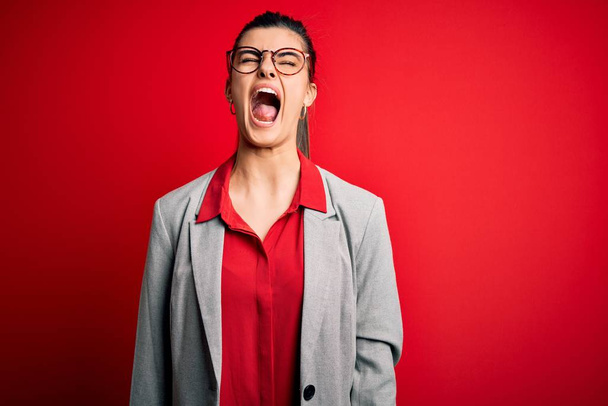 Young beautiful brunette businesswoman wearing jacket and glasses over red background angry and mad screaming frustrated and furious, shouting with anger. Rage and aggressive concept. - Фото, изображение